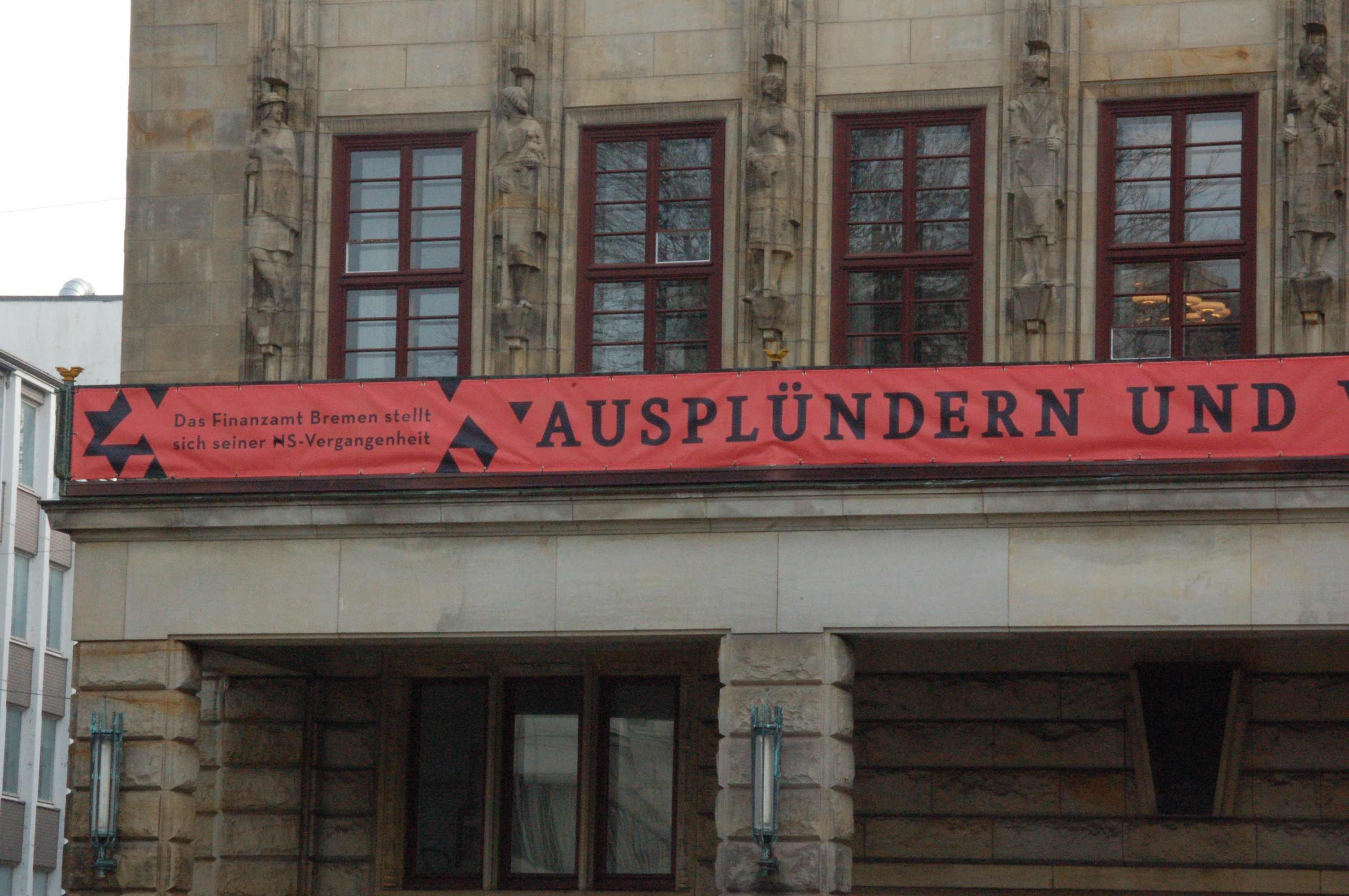 <p>Das Bild zeigt den Haupteingang des Bremer Finanzamts. Über den Türen ist ein roter Banner angebracht mit der Aufschrift: Ausplündern und...-Das Finanzamt Bremen stellt sich seiner NS-Vergangenheit.</p>