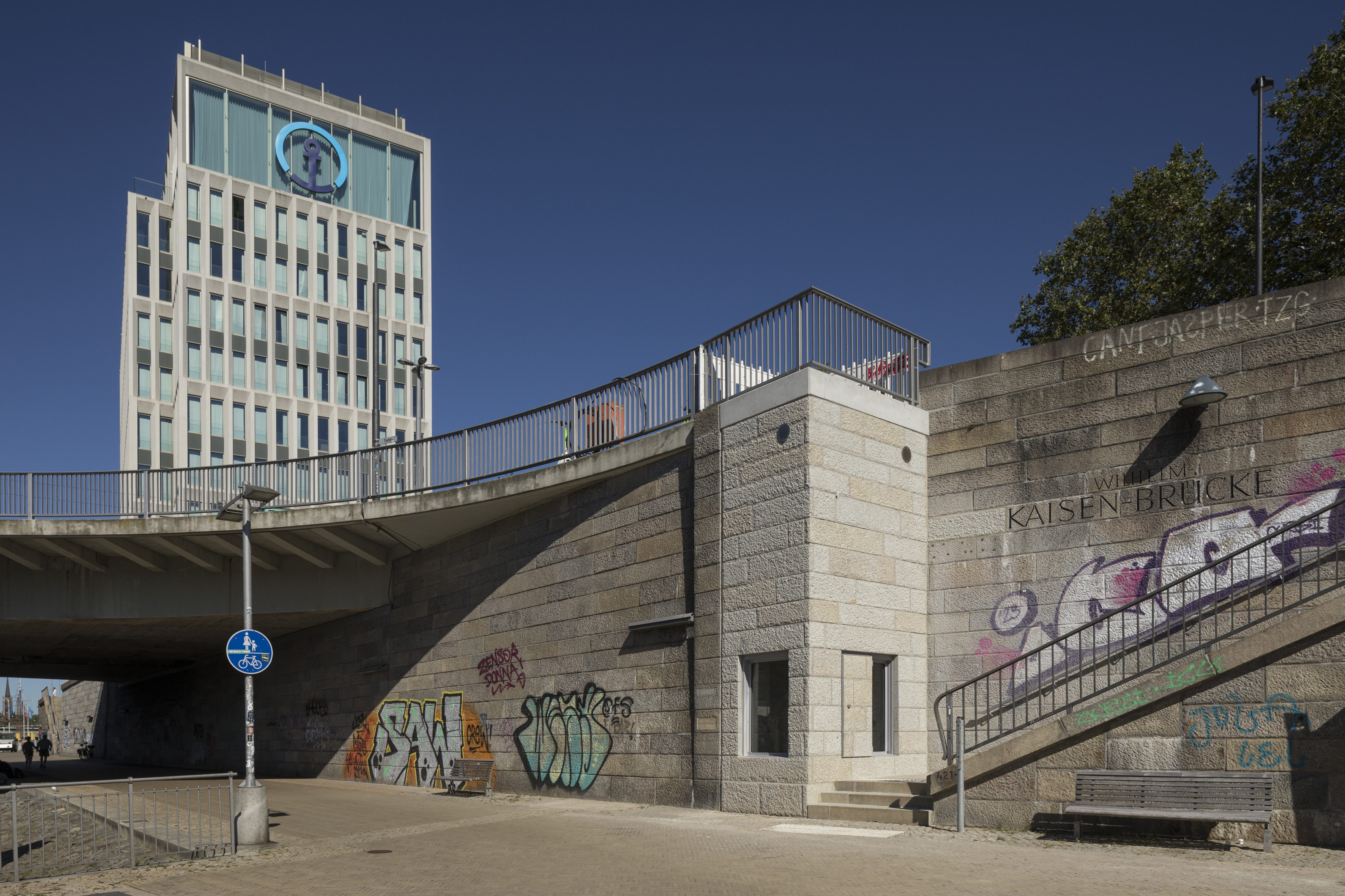 <p>Blauer Himmel. Auf der Wilhelm-Kaisen-Brücke der Neubau des Bürogebäudes von Kühne und Nagel. Unterhalb das fast fertig gestellte Mahnmal. Unlesbare Graffitis an der Wand.</p>