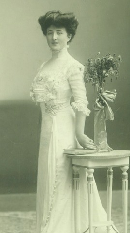 <p>Eine junge Frau steht im weißen Kleid neben einem kleinen Tisch mit Blumen darauf und blickt strahlend in Richtung der Kamera</p>